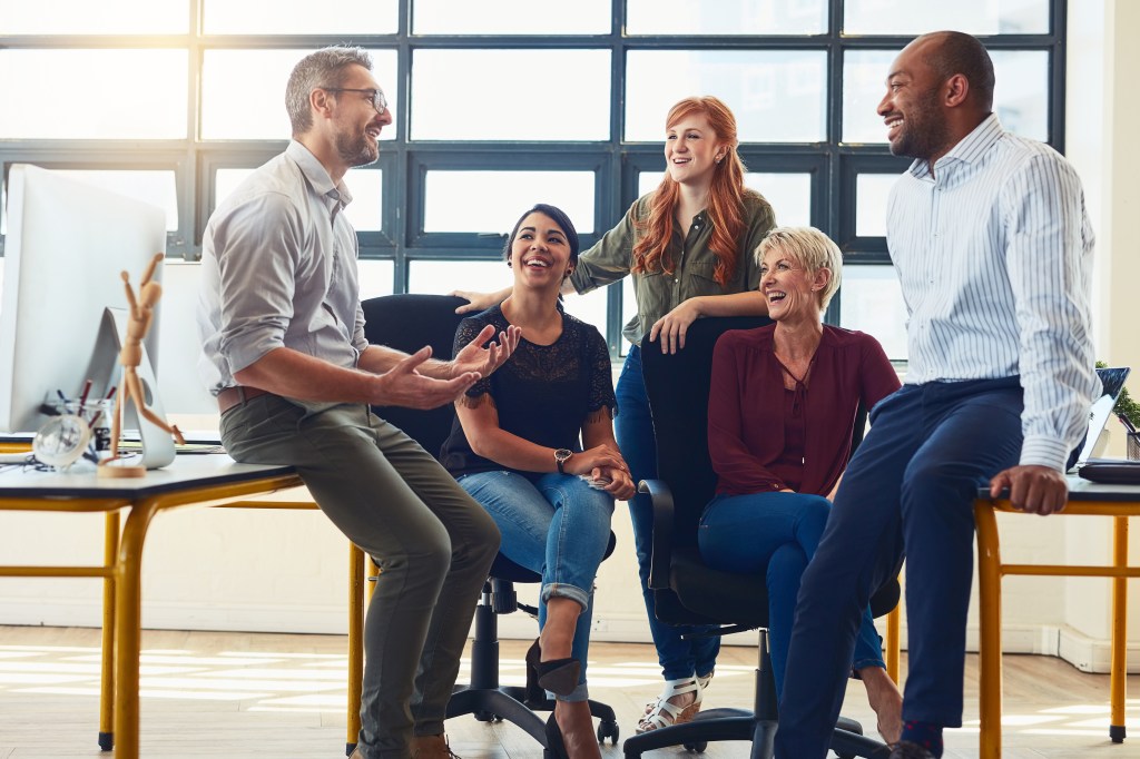 A group of five teammates have a meeting in the middle of their office space while sitting at desks.