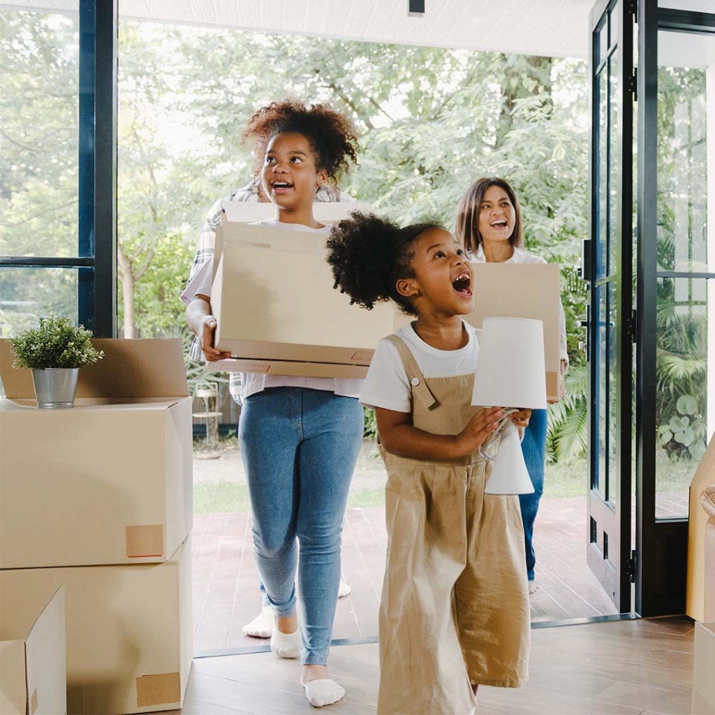 Two children carrying household items walk into their new home followed by two parents.