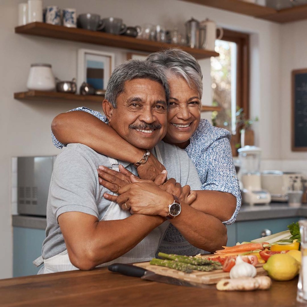 A woman hugs a man from behind while he is chopping vegetables on the kitchen counter.