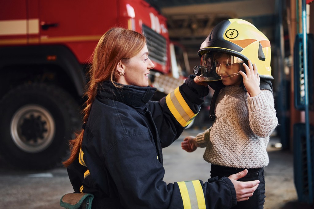 Happy little girl is with female firefighter in protective uniform.