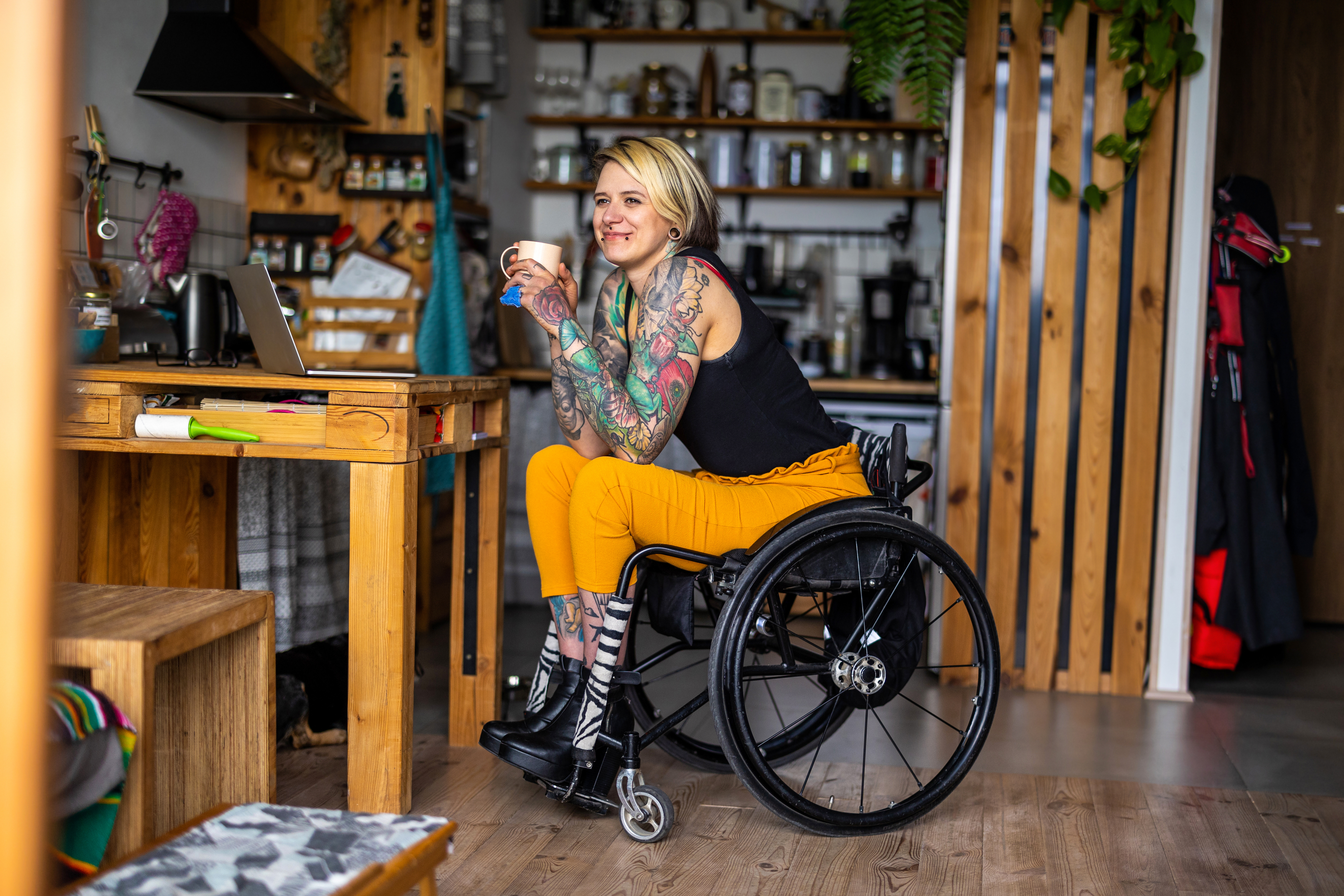 Tattooed female in a wheelchair holds a cup of coffee in her kitchen.