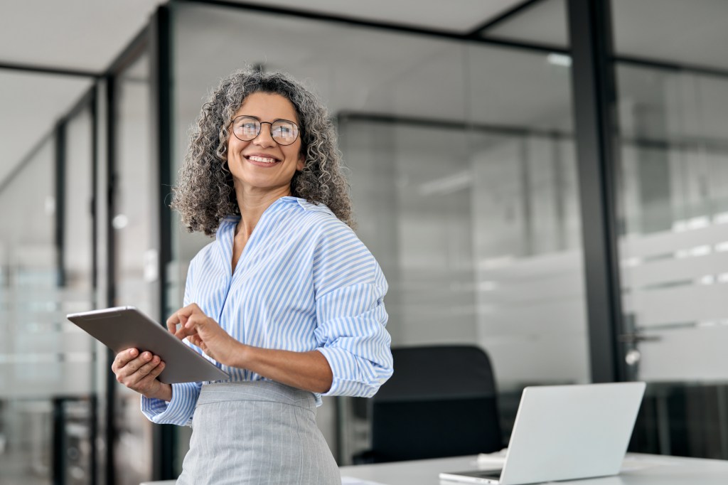 Smiling mature professional business woman holding digital tablet pad standing on office.