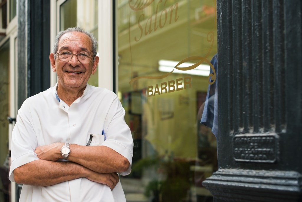 Barber stands in front of barber shop with arms crossed and smiling.