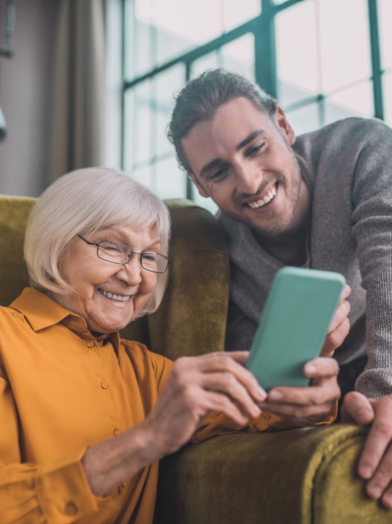 Elderly woman sits in a living room chair looking at mobile device with help from grandson.