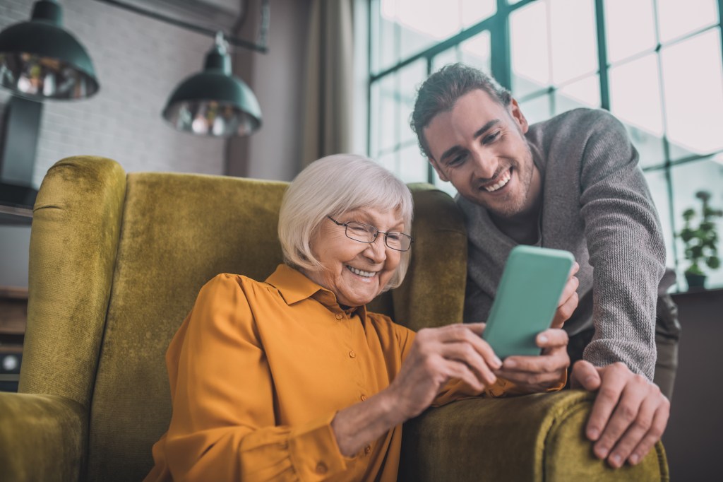 Elderly woman sits in a living room chair looking at mobile device with help from grandson.