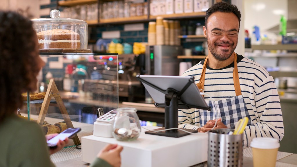 Man With Down Syndrome Serving Female Customer At Coffee Shop Checkout