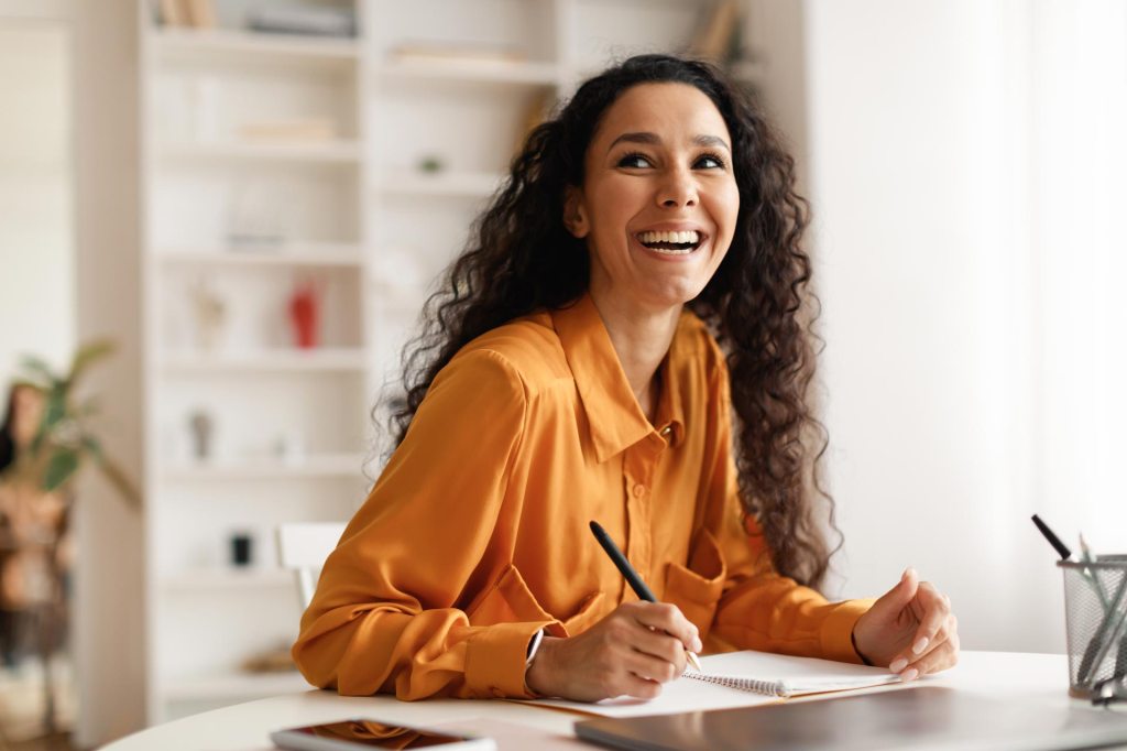 Young female sits at a home desk, journaling in notebook.