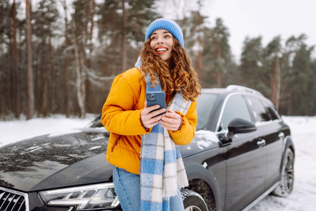 A young woman with a phone stands near her car on a snow-covered road.