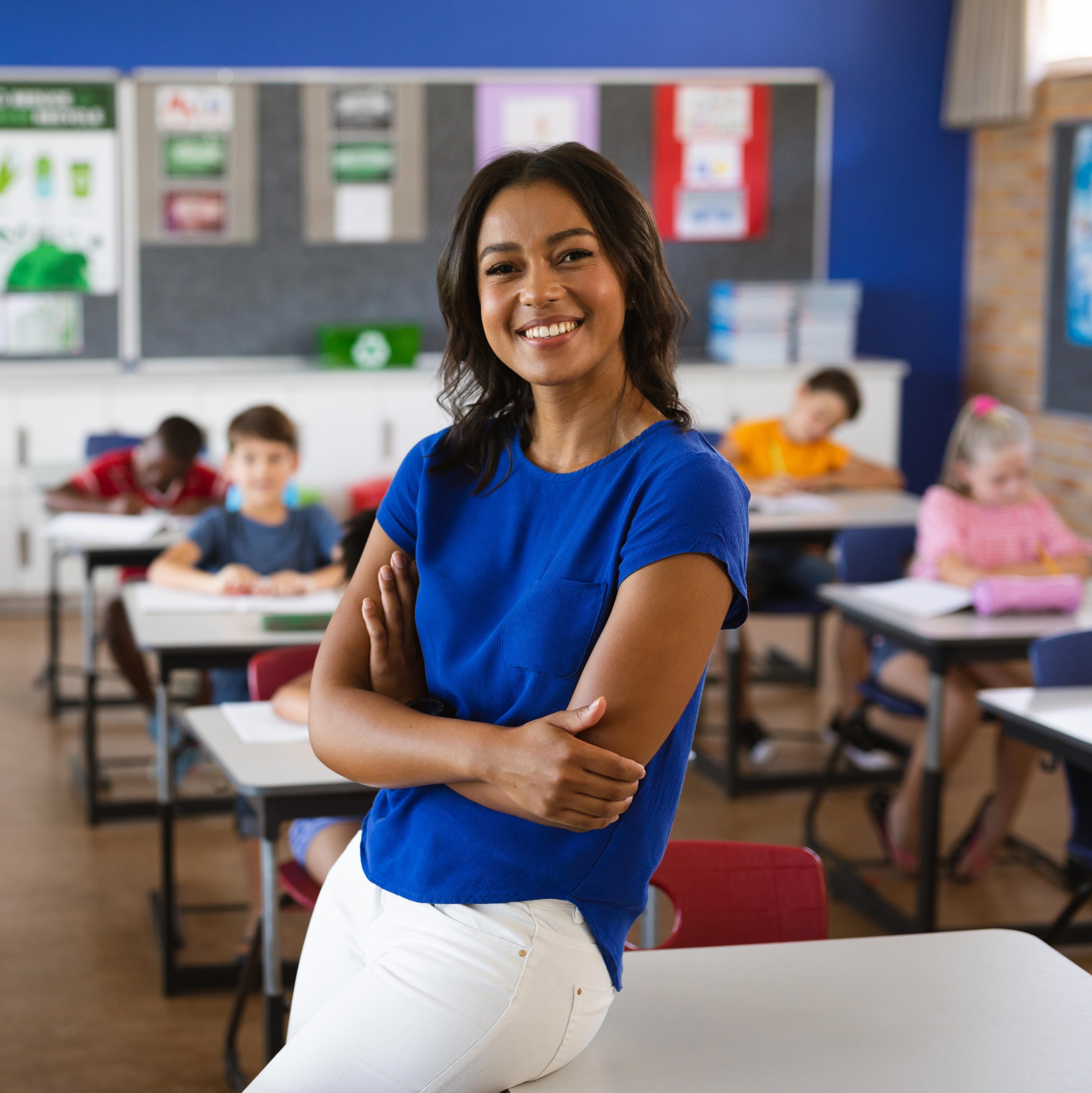 A female teacher sits in on a desk in front of a classroom of elementary school students.