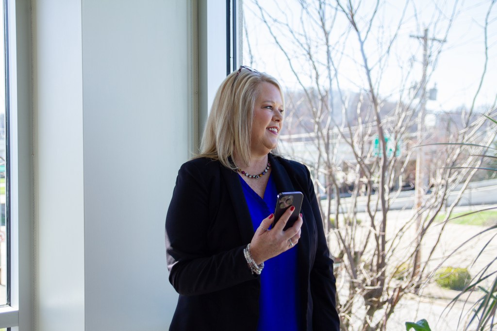 A woman holds a cellphone while looking out bay window onto the city street.