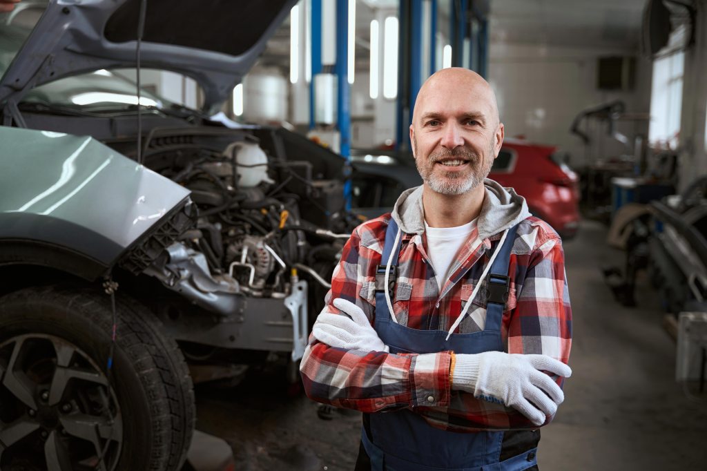 An automotive repair person stands in a three-stall car garage, smiling.